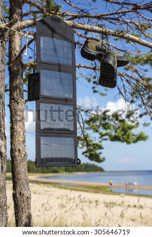 portable solar panel hanging on a pine tree, and charges the mobile phone in the field conditions against the background of the beach with people, near the hanging hiking boots.