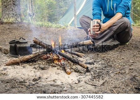 Girl in a blue jacket sitting while camping near the bonfire close to tent is heated and drink hot tea