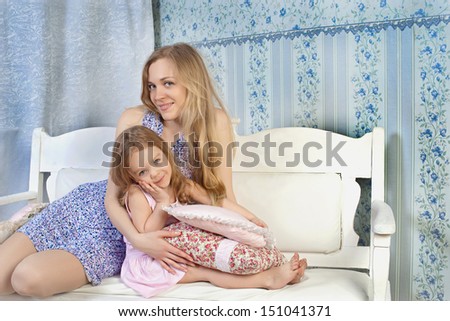 Young mother holding her baby girl in her arms and playing with her while sitting on a white sofa at home, enjoying motherhood and having fun.