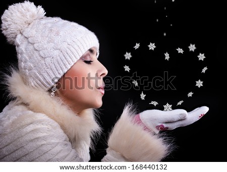 Girl in a fur coat, hat, and gloves on a black background blows away with the palms snowflakes