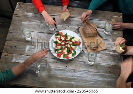 Eating caprese salad with friends, top view on rustic table with hands, bread and big plate with mozzarella, tomatoes and basil leaves (also available as footage)