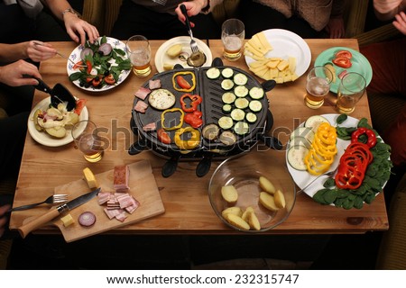Raclette grill party friends eating grilled vegetable drinking beer on vintage wooden desk Raclette grill party friends eating grilled vegetable drinking beer on vintage wooden desk