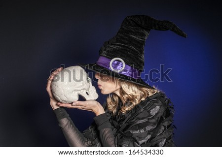 pretty woman dressed as Halloween witch kissing a skull isolated on dark blue background
