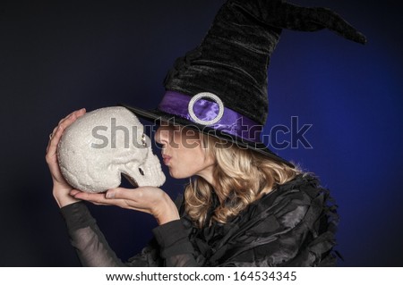 pretty woman dressed as Halloween witch kissing a skull isolated on dark blue background