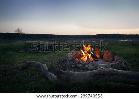 Campfire at dusk with woodpile and lake in background