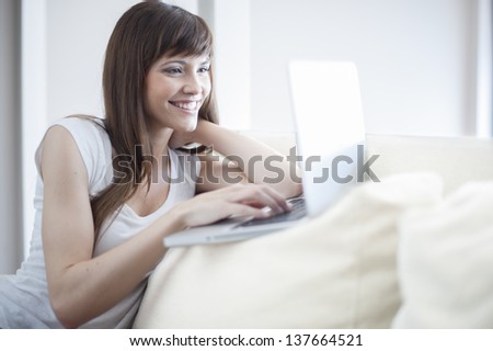 Smiling woman using her laptop sitting on the couch in her home. She is looking at laptop and smiling