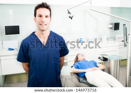 Dentist in his surgery. In the background we can see the patient. He poses looking at camera with arms behind back