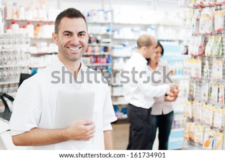 Portrait of a pharmacist holding a tablet. In the background we can see two customers at the pharmacy
