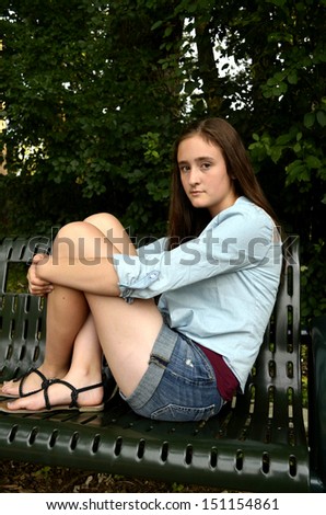 Portrait of female teen sitting on green park bench with green leaf background hugging knees with serious expression