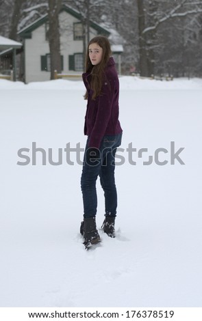 Teen-aged girl walking away in the snow looking back with irritated expression wearing plum pea coat, jeans, and black combat boots