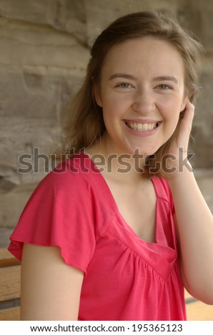 Portrait from the chest up of a pretty young caucasian woman with blond hair and green eyes smiling with hand tucking hair wearing a pink spring top on rustic background
