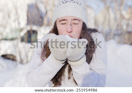 Pretty brunette girl outside in the snow  wearing white knit hat and mittens blowing handful of snow with eyes closed