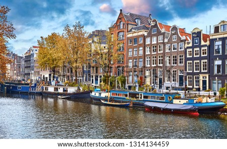 Amsterdam, Netherlands. Floating Houses, houseboats and boats at channels by banks. Traditional dutch dancing houses among trees. Evening autumn street above water blue sky with clouds.