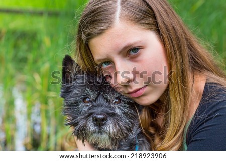 Blonde caucasian teenage girl embracing and hugging black dog in nature with green reed in background
