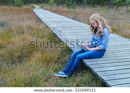 Blonde girl working on tablet computer on wooden path in nature