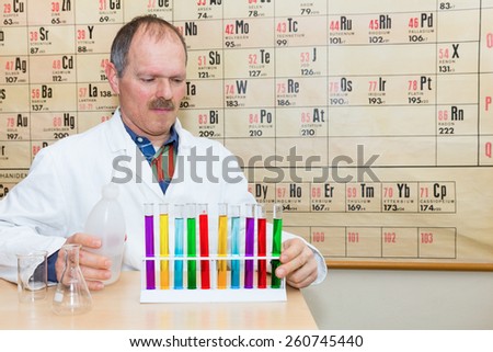 Chemist filling glass test tubes with colored liquids in front of periodic system in classroom