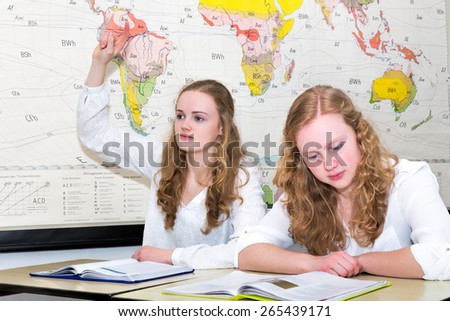 Caucasian teenage girl with learn finger in geography lesson. Two european sisters sitting side by side in high school class. One is reading a textbook to prepare for her study.