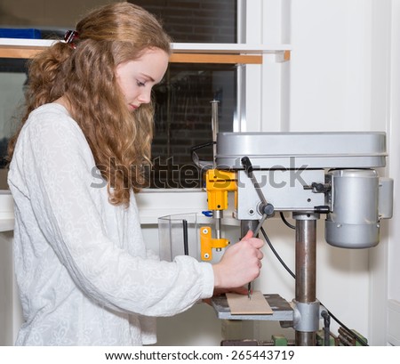 Caucasian teenage girl operating electric drilling machine. The european girl with long hair drills a hole in a piece of wood during technique lesson on high school. learning practice in education