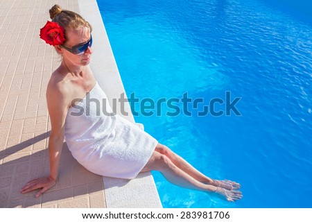 Caucasian woman in white towel with red rose and sunglasses cooling her feet in water of blue swimming pool