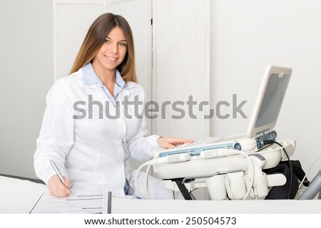 Portrait of a young female radiologist sitting by ultrasound machine while smiles and writes on a medical information form