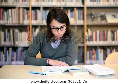 portrait of pretty female girl studying for exam. long brown hair girl with black glasses student in a library with bookshelf on background