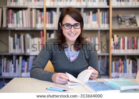 long brown hair girl with black glasses studying and smiling in a library  - portrait of pretty female student at work with books and pen on the desk