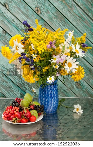 still life bouquet of chamomiles, yellow and blue flowers on the blue vase with berries on the plate
