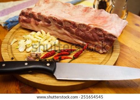 Fresh pork ribs, meat marinated and prepared for roast with garlic. Laying on a wooden table on a round cutting board with knife, chilly peppers and garlic.