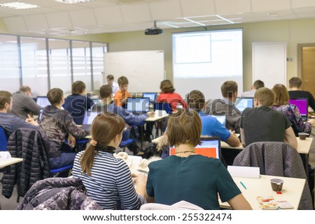 people sitting rear at the computer class at the desks with notebooks and the trainer near the screen explaining the task