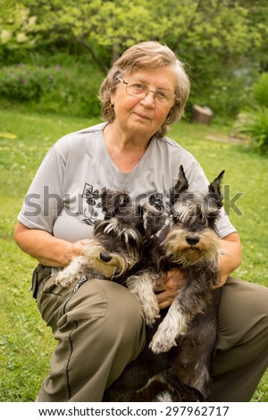 Happy attractive senior woman in glasses sitting with two black and silver miniature schnauzer dogs in a garden