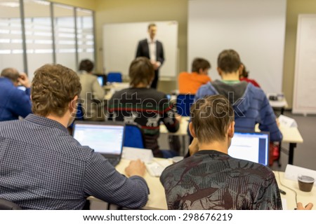people sitting rear at the computer class at the desks with notebooks and the trainer near the screen explaining the task