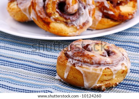 one cinnamon roll with plate of cinnamon rolls behind it wit depth of field focus on front cinnamon roll