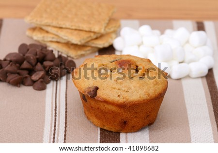 Smores muffin with chocolate chips, graham crackers and mini marshmallows on a brown striped place mat