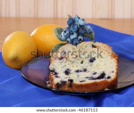 Blueberry lemon bread slices on a blue plate with artificial lemons and blueberries as accents on blue satin fabric