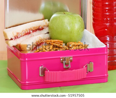 pink metal lunchbox with peanut butter and jelly sandwich, pretzels, apple and red drink on a green placemat close up horizontal composition