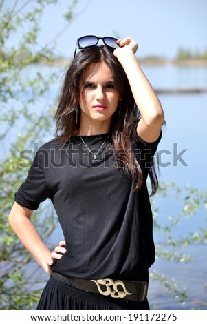 Beautiful young girl in black dress and sunglasses on the background of nature and water