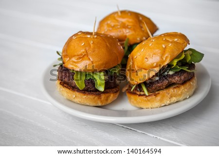 Mini Beef Sliders With Arugula On Sesame Seed Buns On A White Washed Wood Background