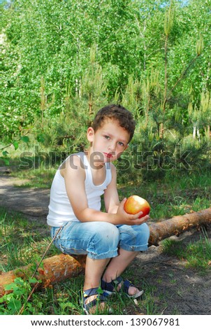 In the summer, bright sunny day, the boy sits in a pine forest on a log and eats an apple