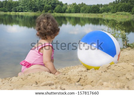 In the summer, on the beach near the lake in the sand little girl playing with a ball
