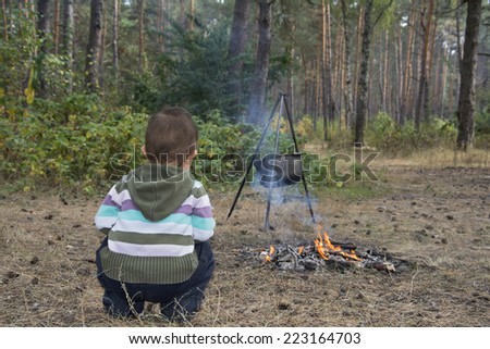 A small child in a pine autumn forest sits near the fire where the food is cooked in a cauldron.