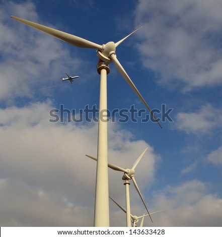 Perspective image from below of aeolian park with towering wind turbines and aircraft flying above, on blue sky and clouds
