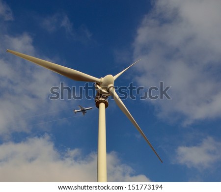 Perspective image from below of towering wind turbine and aircraft flying above, on blue sky with clouds background