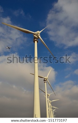 Vertical perspective from below of wind farm with towering mills and aircraft flying above, on blue sky and clouds