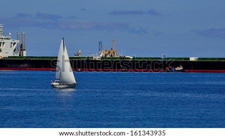 Small sailboat sailing on the calm sea and next to a large tanker docked