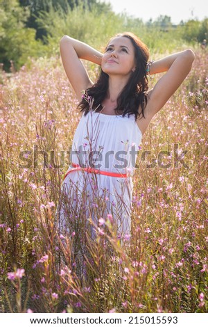 Young brunette woman in a white dress. A girl stands in the middle of pink flowers field on a sunny day. Field, flowers beauty, nature,  - The concept of country vacation. Article about vacation.