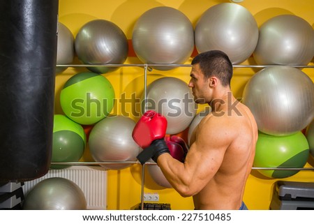 Muscular man in the gym. The man in red boxing gloves, hit a punching bag, exercise. Boxing, workout, muscle, strength, power - the concept of strength training and boxing