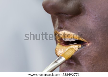 Putting drawing on the model\'s face. Makeup artist draws golden lips on  face of the model with a brush.Golden lips on dark make-up face, creativity, make-up, the idea, brush, creative process.