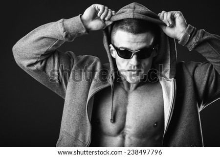 Young muscular man with open jacket revealing muscular chest and abs.Portrait of a muscular young man in hood jacket posing on black background.Young man with athletic body posing on black background
