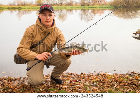 A young man with a fish in his hands and spinning. A fisherman on the river caught zander. Man fisherman sat on his legs and holding a fish in his hands.