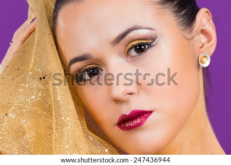 Girl with a professional makeup posing on a purple background. Beauty photo. Gold cloth near the girl\'s face. gold fabric, cloth. Makeup executed in gold style.The perfect make-up lips and eyes